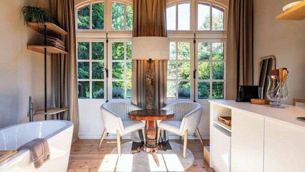Une salle de bain avec une baignoire, deux chaises, une table ronde et une fenêtre avec des rideaux.