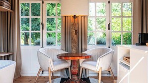 Une pièce comprenant une table ronde en bois, deux fauteuils rembourrés, une grande fenêtre avec des rideaux et une bibliothèque.