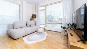 Ein modernes Wohnzimmer mit grauem Sofa, einem kleinen runden Tisch, einem großen Fenster mit Vorhängen, einer Stehlampe, einem Fernseher auf einem Holzgestell und einer Pflanze.