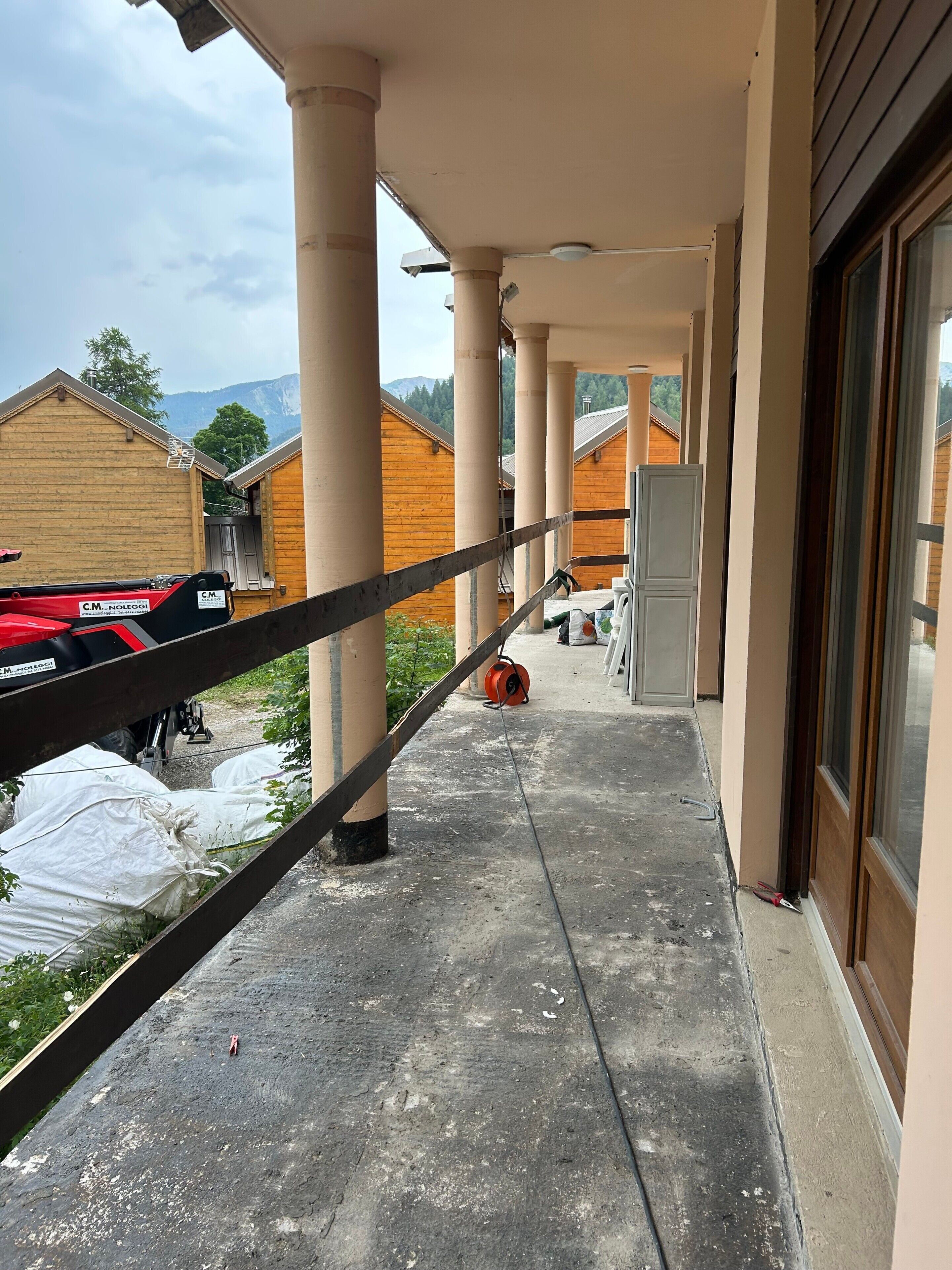Un balcon offrant une vue sur des bâtiments en bois et des montagnes, soutenu par des colonnes, et un véhicule rouge garé à proximité.