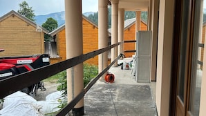 Un balcón con vistas a edificios de madera y montañas, sostenido por columnas, y un vehículo rojo estacionado cerca.