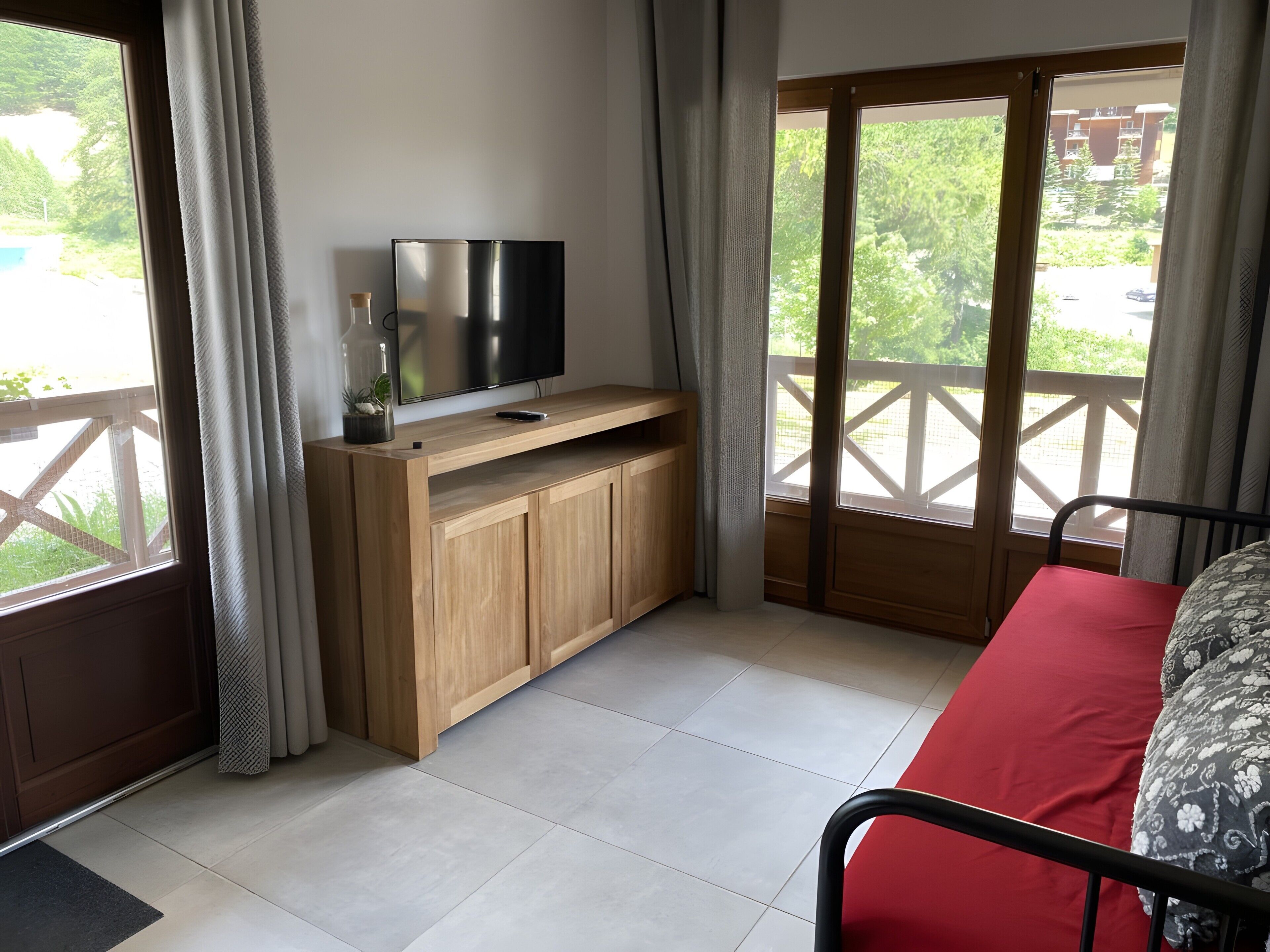 A room with a TV on a wooden cabinet, a bed with red bedding, and a balcony with a view of greenery and buildings.