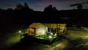 A geodesic dome tent with a platform, illuminated by internal lighting, surrounded by a fenced area with a wooden deck and a vehicle in the background. - Cozy glamping  in heart of National park Durmitor with private HOT TUBE (Žabljak)