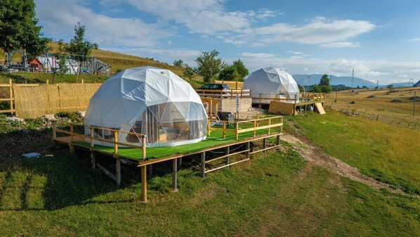 Deux tentes en forme de dôme, dotées d’une terrasse en bois à l’avant, sont installées dans un champ verdoyant sous un ciel dégagé.
