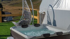 Un jacuzzi con una silla colgante, rodeado por una carpa blanca y una terraza de madera, sobre un paisaje cubierto de hierba.