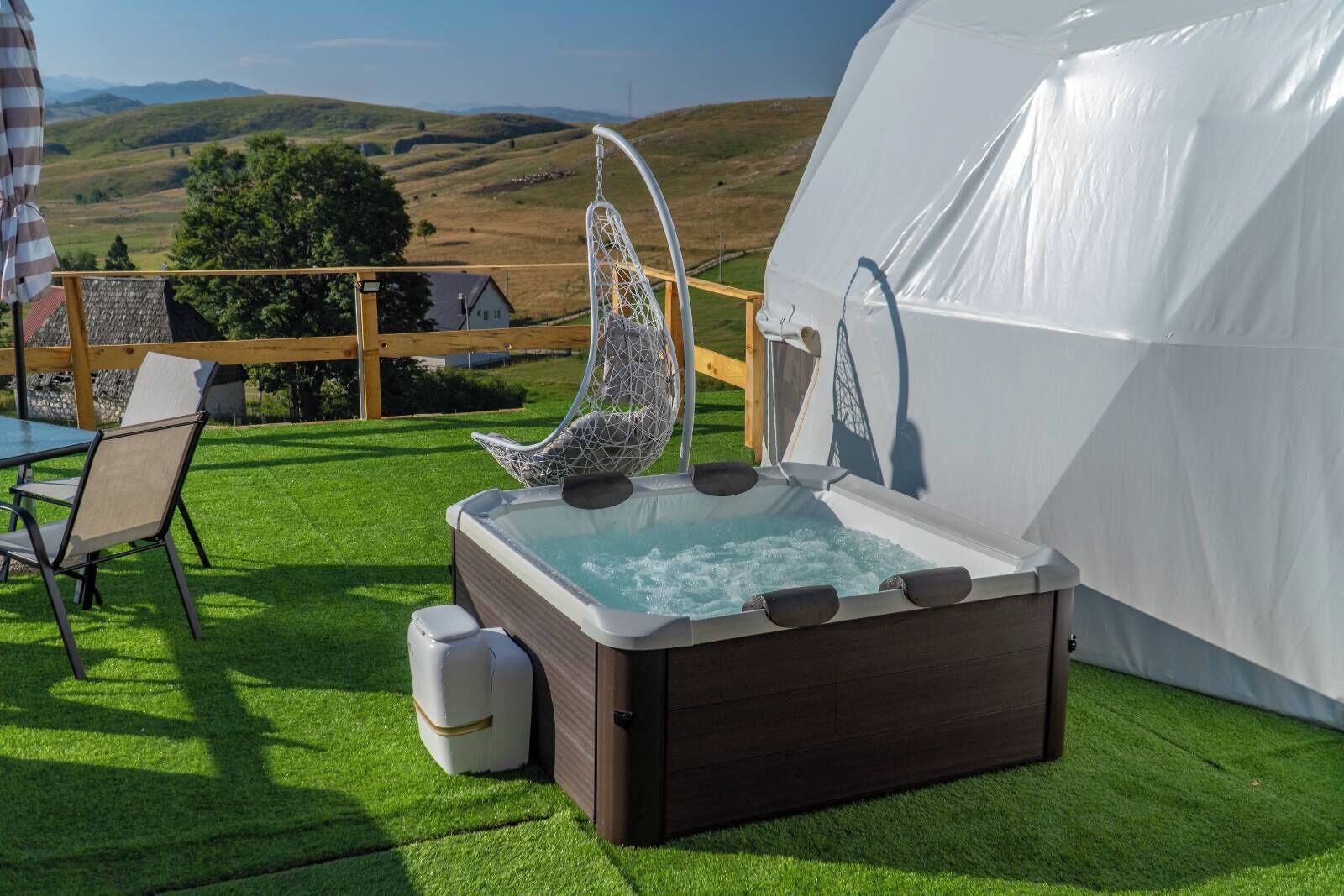 A hot tub with a wooden frame, a hanging chair, and a table with chairs on a grassy lawn with a view of hills.