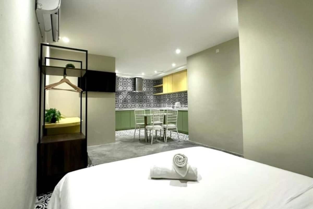 Una camera d'albergo con un letto rifatto, una piccola area salotto con un tavolo e sedie, un angolo cottura con piastrelle decorate e un televisore montato a parete.