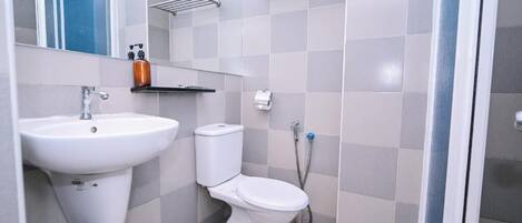 Et badeværelse med en hvid vask, toilet og bruser. Der er hylder over toilettet og et spejl over vasken.