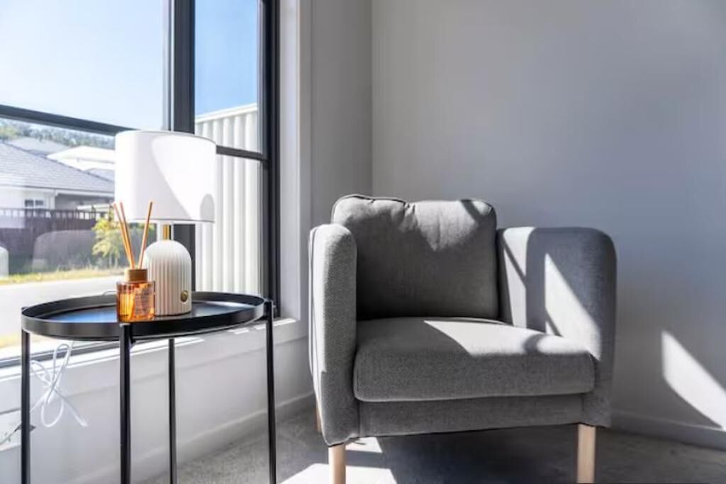 A corner room with a grey armchair, a small round table with a lamp, and a view of a building outside.