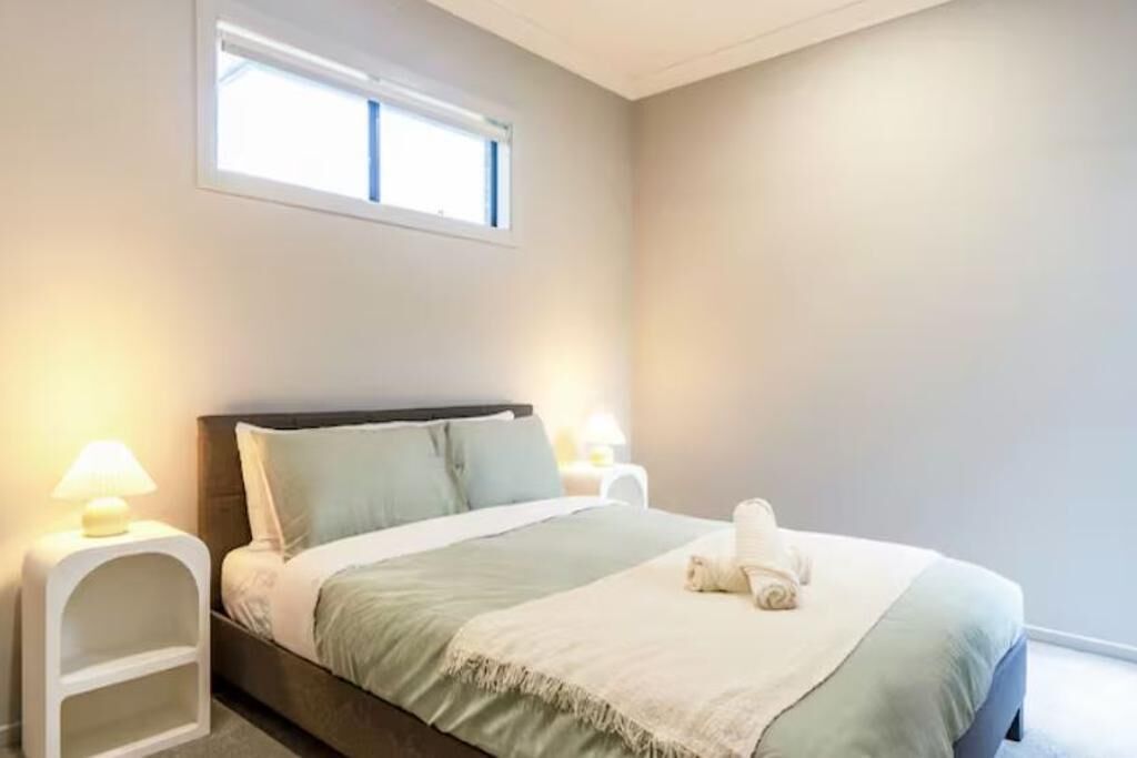 Una cama bien hecha con una colcha verde y blanca, dos lámparas de noche y una ventana sobre la cama.