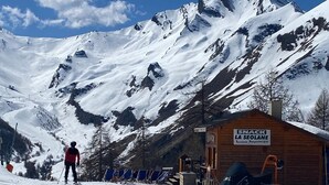 Ein mit Schnee bedeckter Berg mit einem Holzhaus, auf dem 'SNACK LA SEOLANE' steht, und einer Person beim Skifahren.