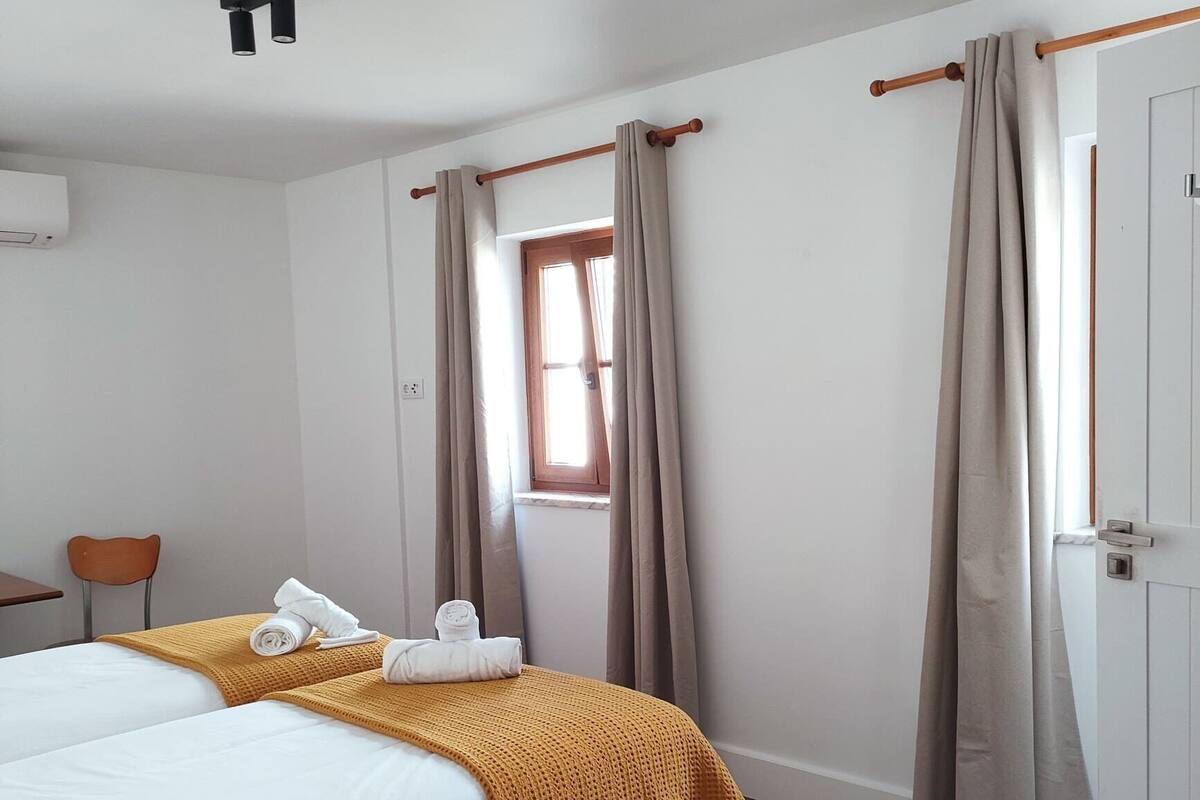 A hotel room with two beds, a wooden floor, a window with curtains, and a door.