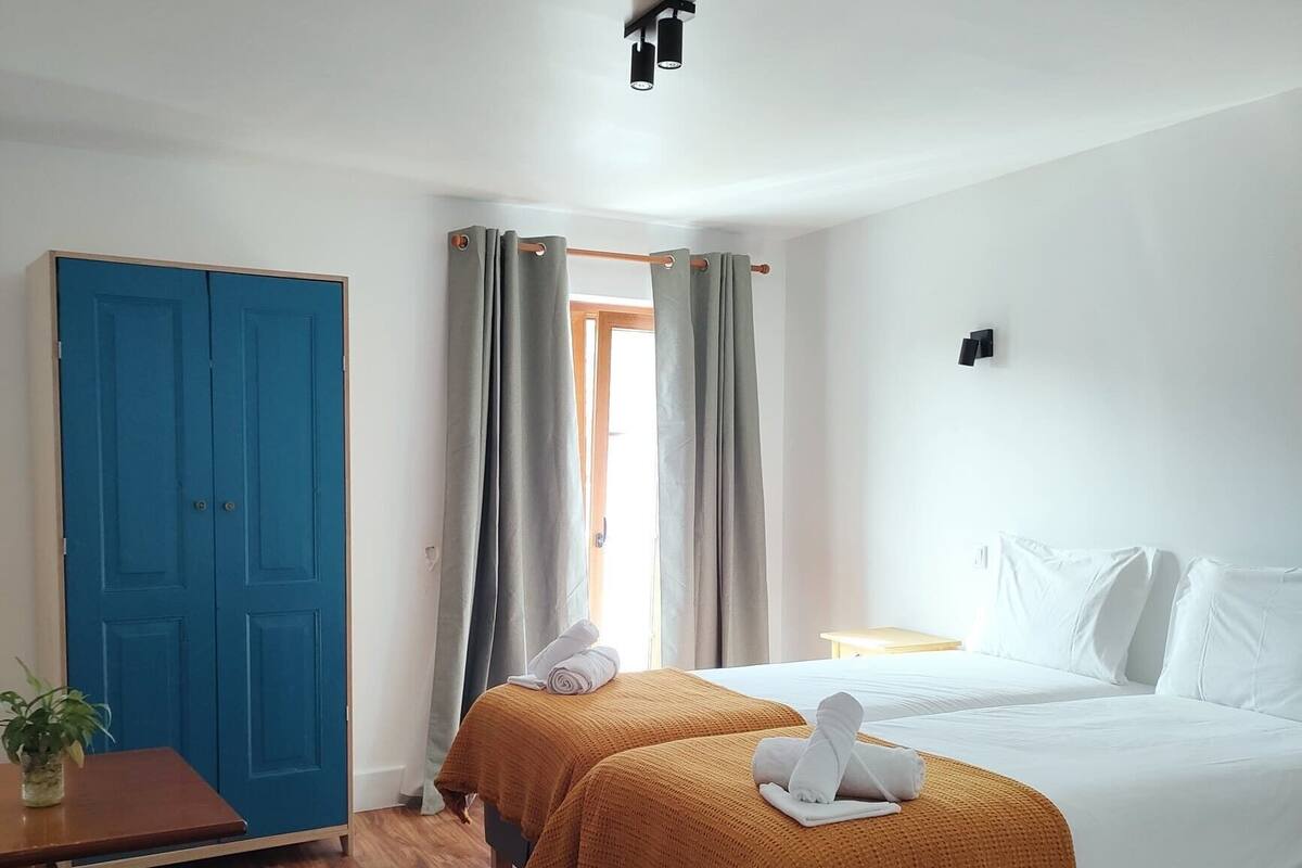 A hotel room with two beds, a desk, a blue wardrobe, and a window with curtains.