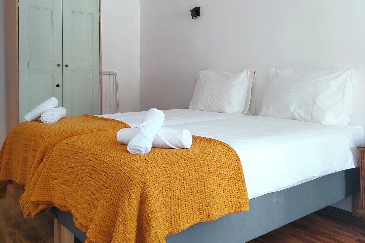 A neatly made bed with white linens and a mustard-colored throw blanket, a wooden wardrobe, a small bedside table, and a wall-mounted light fixture.