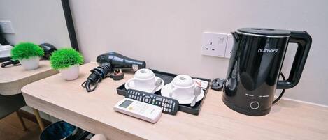 Et hotelværelse med et træskrivebord, en sort kaffemaskine, en vægt, to hvide kopper og to små krukker med planter.
