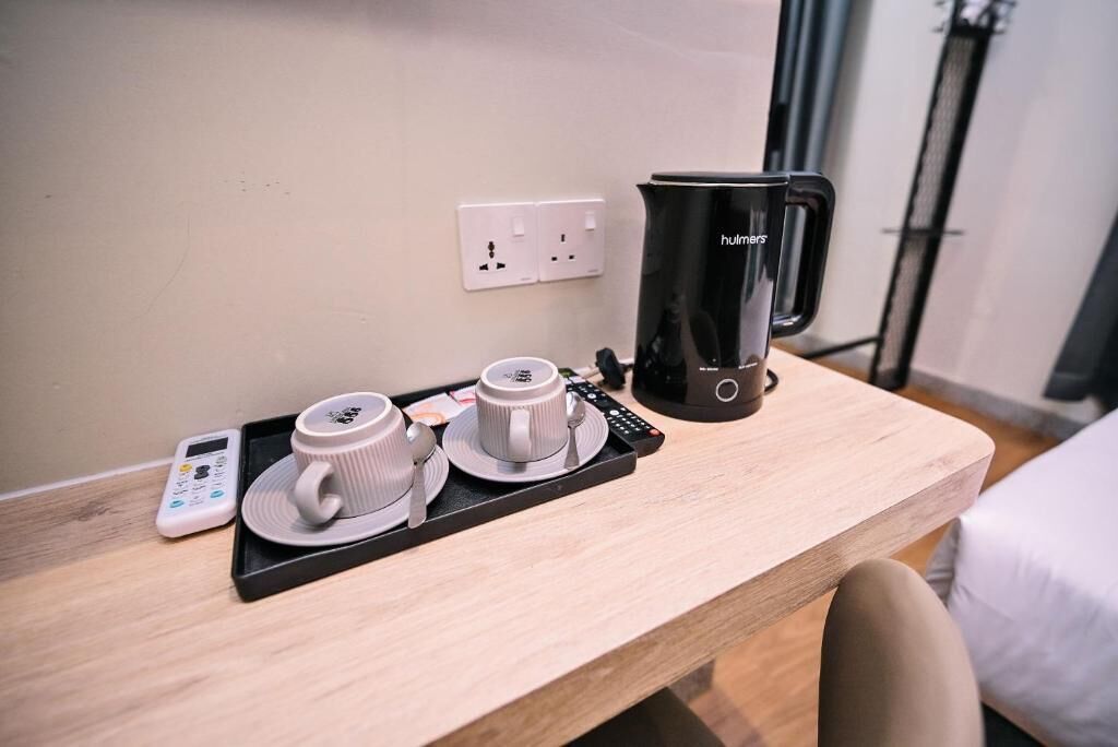 Habitación de hotel con un escritorio de madera, una cafetera negra, dos tazas blancas, una bandeja y un termómetro digital.
