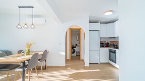 Een moderne keuken met witte kasten, een ingebouwde oven en een magnetron. Er is een eethoek met een houten tafel en stoelen, en een gang die leidt naar een wasruimte.