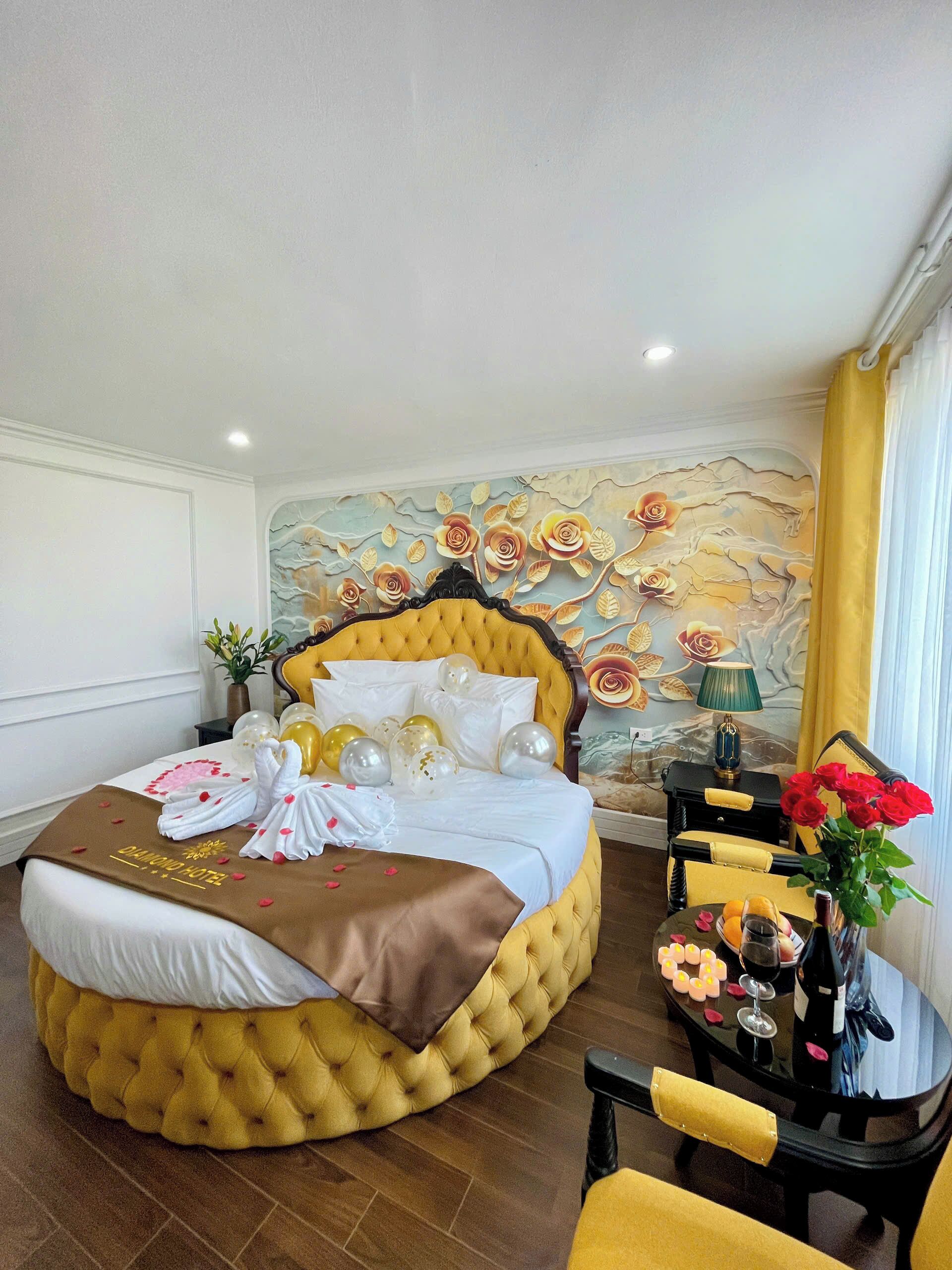 Une chambre d’hôtel avec un lit jaune à motifs capitonnés, du linge de lit blanc, une fresque murale décorative, une petite table surmontée d’un vase de roses rouges et une chaise.
