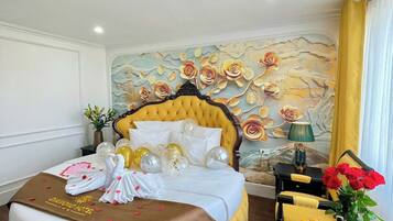 Une chambre d’hôtel avec un lit jaune à motifs capitonnés, du linge de lit blanc, une fresque murale décorative, une petite table surmontée d’un vase de roses rouges et une chaise.