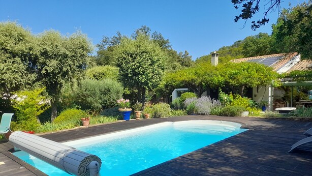 A swimming pool with a wooden deck, surrounded by lush greenery and a small stone structure. - Beautiful modern villa with swimming pool in the heart of the garrigue (Lauret)