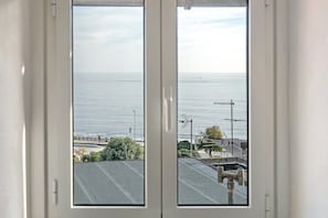 Uitzicht door een raam, met blauwe lucht en een rustige zee, een straat met een zebrapad en wat groen.