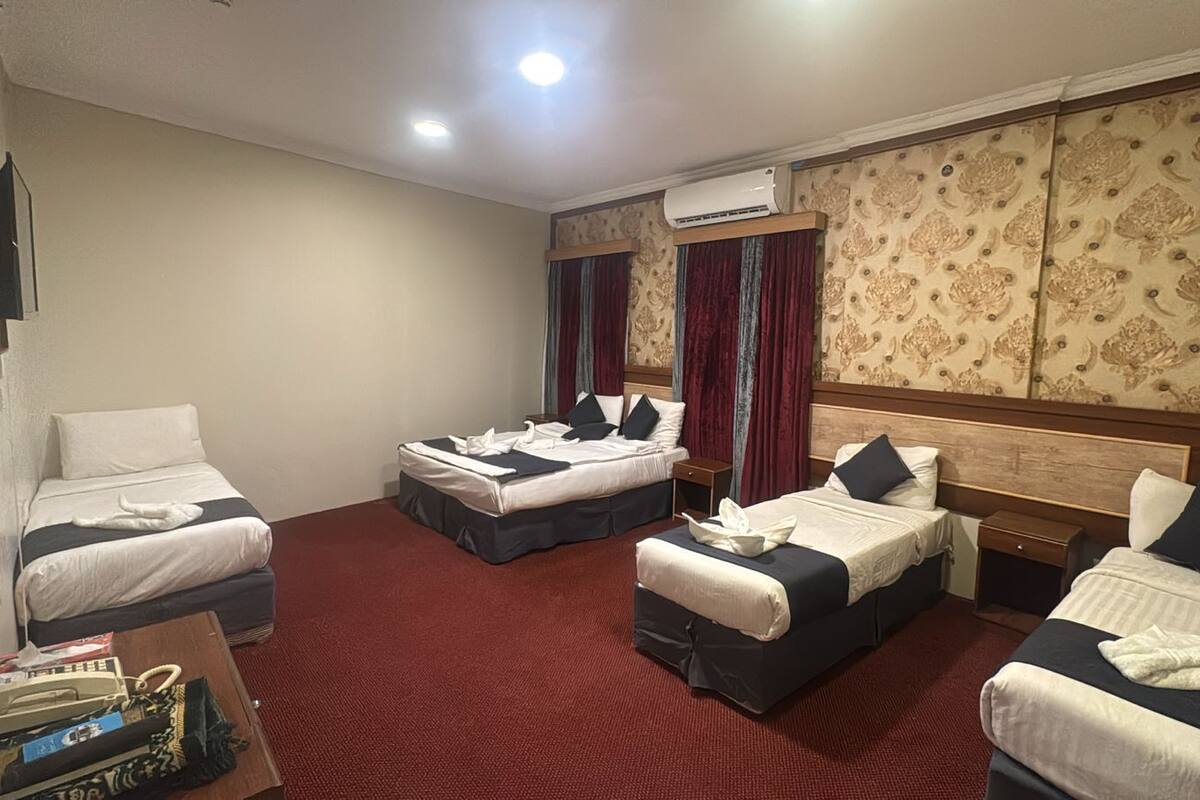 A hotel room with two single beds, a double bed, a wooden headboard, a red carpet, an air conditioning unit, and a small table with a book and a bag.