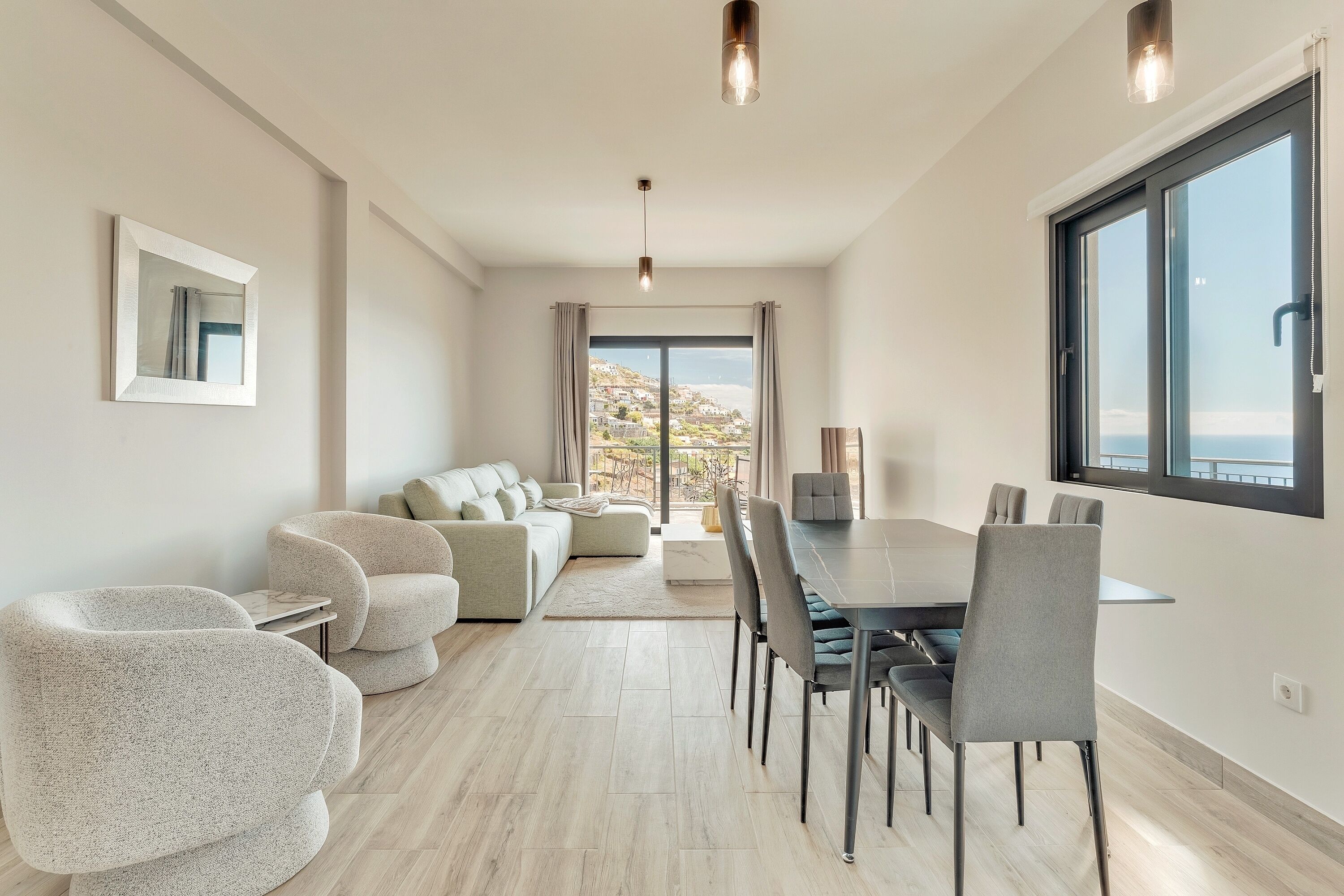 Ein modernes Wohnzimmer mit einer Sofagarnitur, einem Esstisch und Stühlen. Ein großes Fenster bietet Blick auf eine hügelige Landschaft.