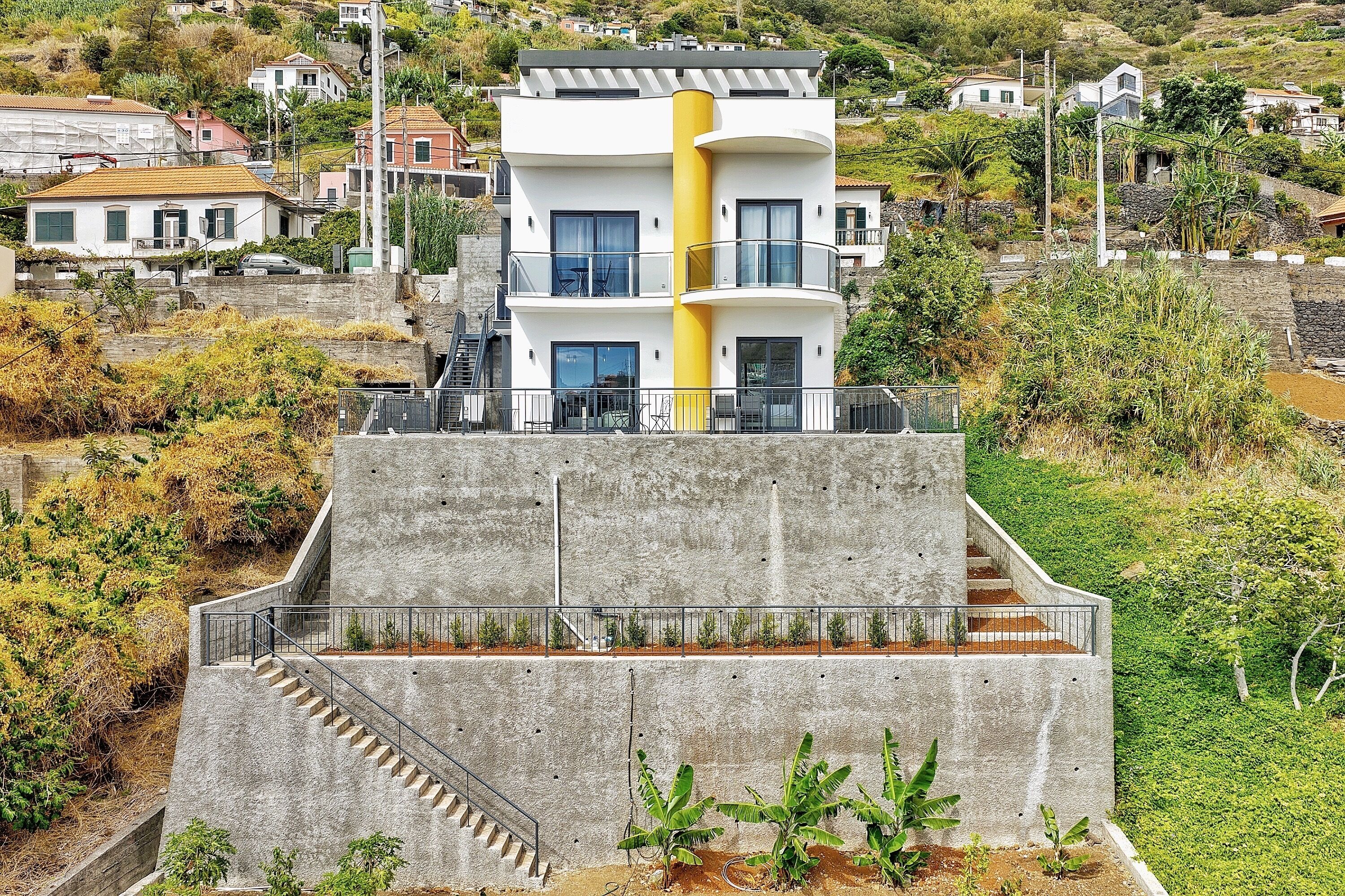 A modern multi-story building with a central yellow column, a concrete staircase, and a surrounding garden.