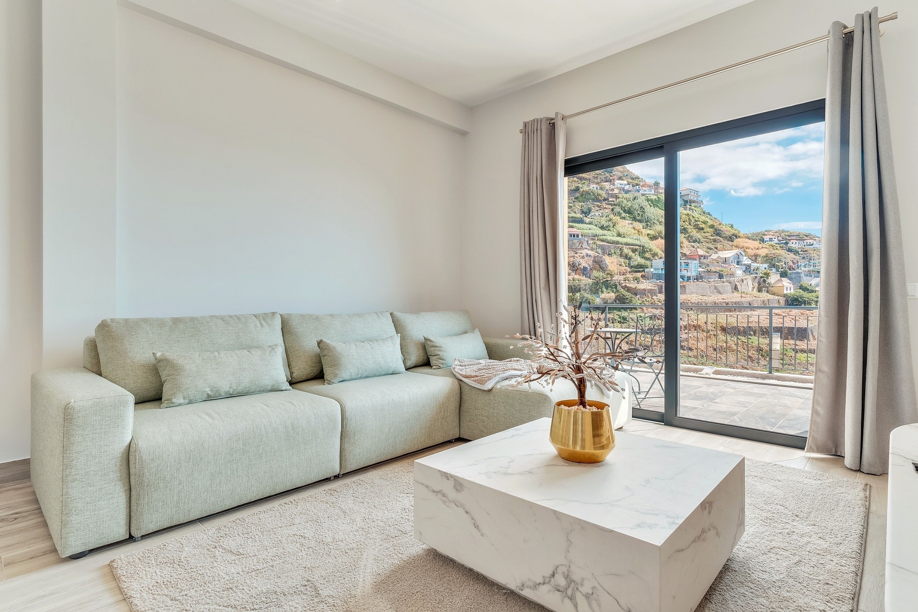 A modern living room with a sofa, a marble coffee table, and a view of a hillside through large windows.