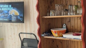 A dining area with a wooden table and chairs, a television mounted on the wall, and a wooden cabinet with shelves containing glassware and plates. - Ground-floor Studio Cabin with terrace. Pets welcome! (leucate)