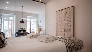 A modern hotel room with a large bed, a desk with a chair, and a view of the city through large windows. - ✨Designer Loft with Balcony in Historic Baixa (Lisbon)
