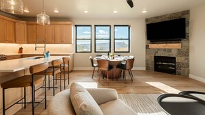 A modern kitchen with wooden cabinets, a bar area, a dining table with chairs, a fireplace, and a TV mounted on a stone wall.