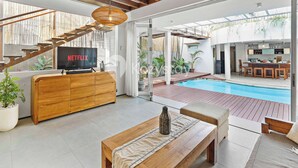 A modern living room with a wooden TV stand, a sofa, and a pool area visible through glass doors. - Ombak Villa by Kozystay - Canggu (Canggu)