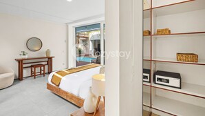A modern hotel room with a bed, a desk, a chair, a lamp, a shelf with baskets, and a view of a pool. - Ombak Villa by Kozystay - Canggu (Canggu)