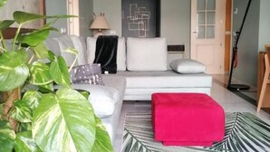 A modern living room with a white sofa, a red ottoman, and a patterned rug. There is a plant in the foreground, a lamp on a side table, and a geometric wall art piece. - Beach and Golf Design Apartment Marbella (Marbella)