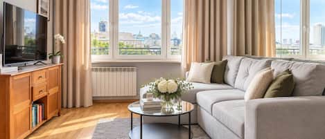 A modern living room with a sofa, a coffee table, a TV, and a view of the cityscape through large windows.