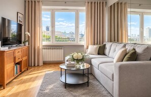 Ein modernes Wohnzimmer mit einer Couch, einem Couchtisch, einem Fernseher und Blick auf die Stadt durch große Fenster.