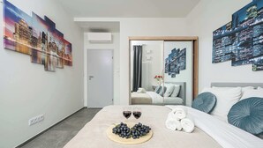 A hotel room with a bed, a tray of grapes, two glasses of red wine, and a cityscape painting on the wall. - 2-Room Apart on 15th Floor (Warszawa)