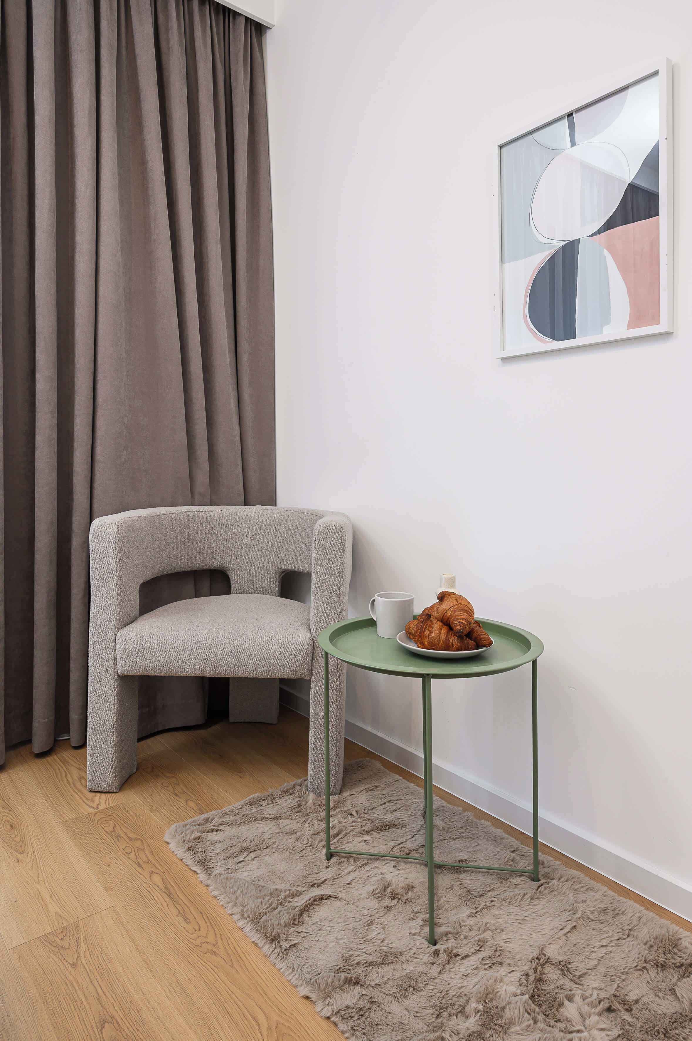 A room with a grey upholstered chair, a small round table with a plate of croissants and a cup, a fluffy grey rug, and a framed abstract painting on the wall.