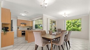 A modern kitchen with a dining area, featuring a large island, wooden cabinets, and a view of greenery through the windows. - 3 minute drive to the beach! Family home with a BBQ, boardgames, and a backyard. (Callala Bay)