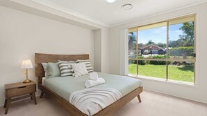 A bedroom with a large window, a wooden bed, a nightstand with a lamp, and a view of the outside. - 3 minute drive to the beach! Family home with a BBQ, boardgames, and a backyard. (Callala Bay)
