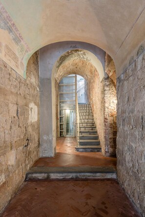 Un couloir voûté aux murs de pierre, un escalier en bois et une rampe en métal.