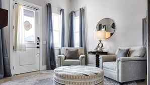 Ein Wohnzimmer mit einem runden Hocker, zwei Sesseln, einer Lampe und einem Spiegel an der Wand.