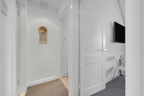 A hallway with white walls, a round mirror with fringe, a flat-screen TV mounted on the wall, and a folding chair.