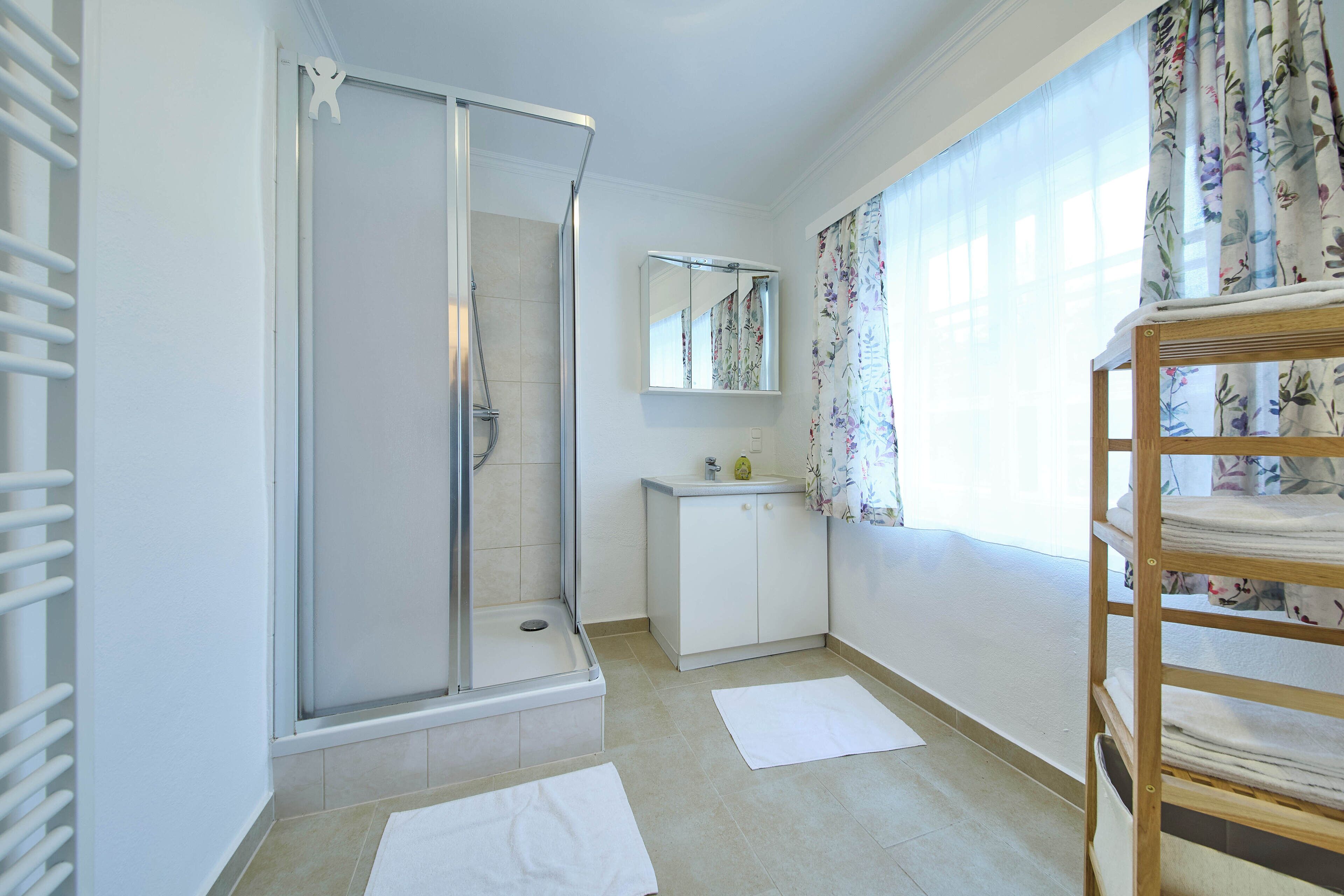 A bathroom with a shower, a sink, a mirror, and a window with floral curtains.