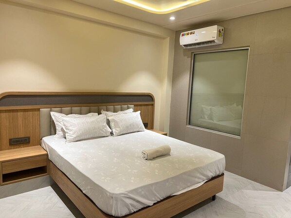 Una cama bien hecha con edredón blanco y almohadas, cabecera de madera, mesita de noche y un equipo de aire acondicionado montado en la pared.
