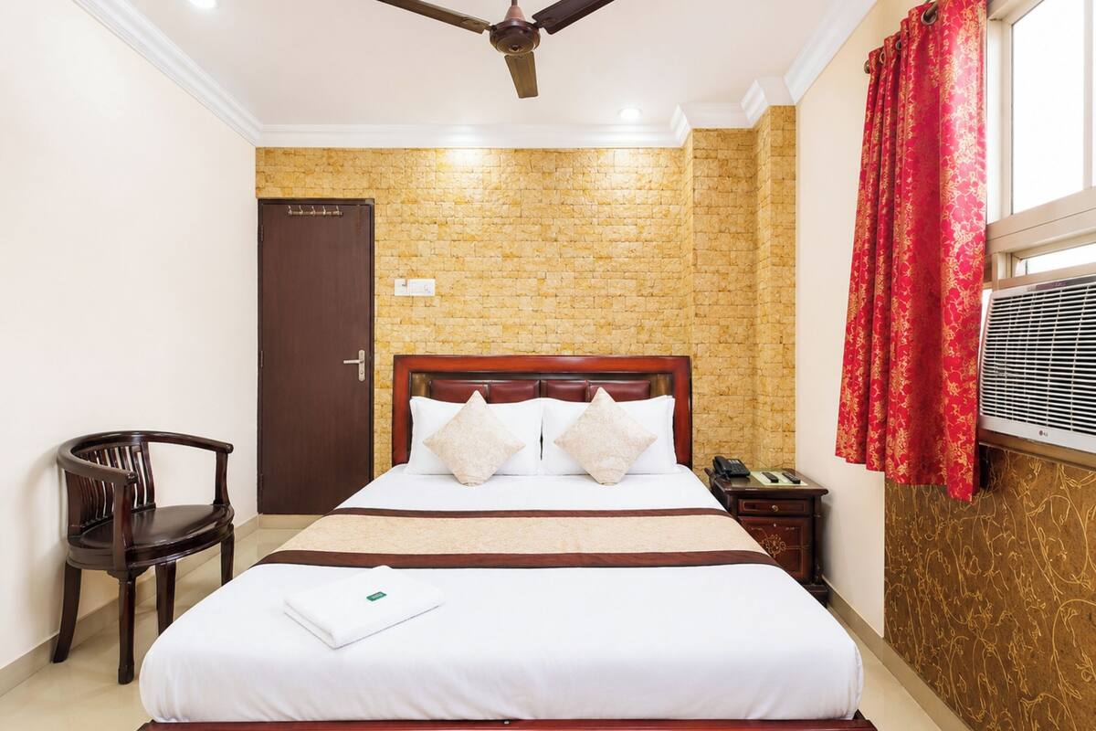 Une chambre d’hôtel avec un mur décoré d’un motif en briques, un lit en bois recouvert de draps blancs, un ventilateur de plafond, une table de chevet en bois, une chaise et une fenêtre avec des rideaux rouges.