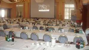 En festlokal med långa bord uppställda för ett formellt evenemang, en stor skärm för presentationer och kristallkronor.