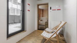 A hallway with a wooden door leading to a sauna room, white tiled walls, and a window with blinds. - Lumina luxury 4-rooms apart with parking (Łódź)