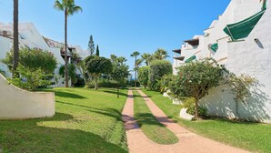 A pathway leading through a well-maintained garden with white buildings, palm trees, and neatly trimmed bushes. - Lunamar Garden first line beach Real de Zaragoza (Marbella)